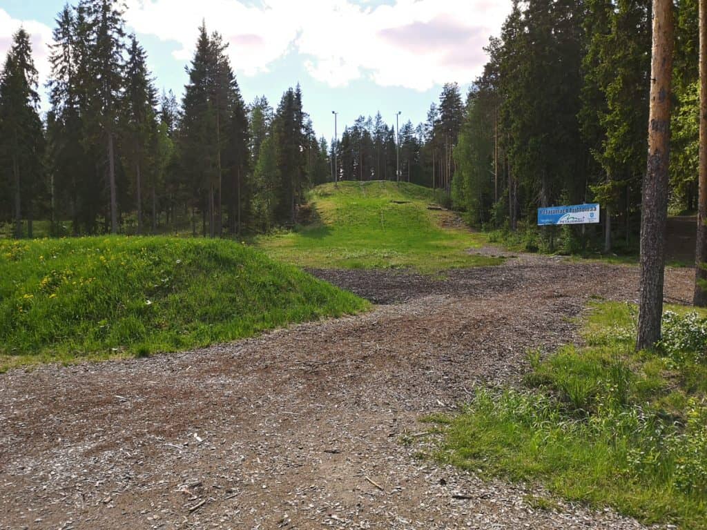 Kuva kesäisestä ulkoilualueesta. Kauempana kyltti "Petäjämäen hiihtomaa". Ympärillä Suomen metsää ja kesäistä luontoa.