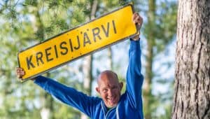 Nauravainen sinipaitainen mies pitelee pään yläpuolellaan keltaista kylttiä, jossa lukee "Kreisijärvi". Taustalla vihreikköä kesäisessä säässä.